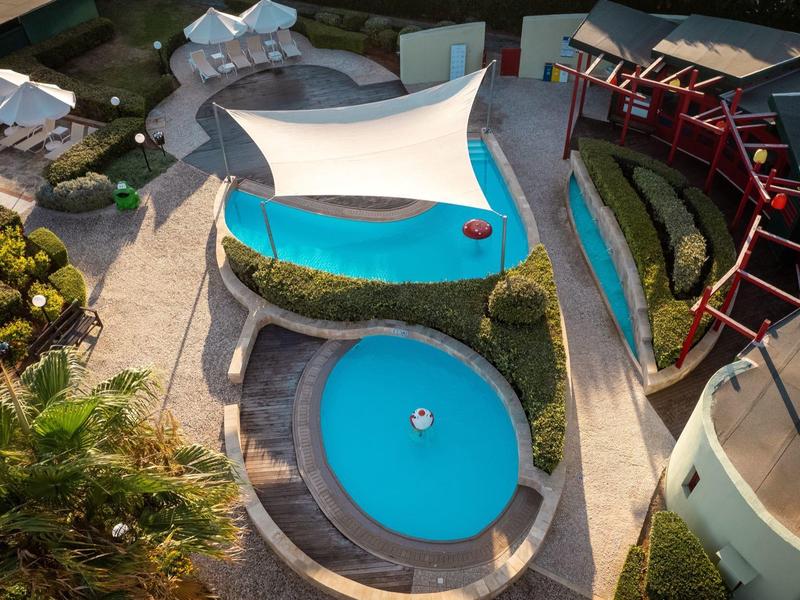 Blick von oben auf zwei runde blaue Pools mit weißem Sonnensegel und grün bepflanzten Wegen.