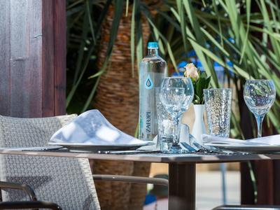 Ein gedeckter Tisch mit Gläsern, Servietten und einer Wasserflasche vor grünen Pflanzen.