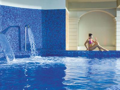 Großer, blauer Pool mit Wasserstrahlen und Frau in pinkem Badeanzug an hellem Wandbogen sitzend.