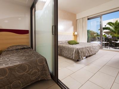 Zimmer mit Einzelbett und Doppelbett, Glaswand dazwischen, Balkon mit Pflanzen im Hintergrund.