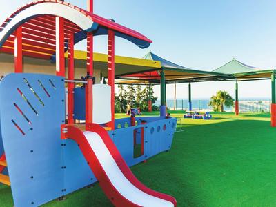 Bunter Spielplatz mit Rutsche, blauem Klettergerüst und schattigen Sitzbereichen auf grünem Boden.
