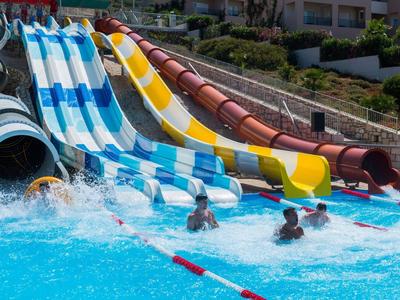 Mehrere bunte Wasserrutschen führen ins blaue Schwimmbecken, wo Kinder ins Wasser springen.