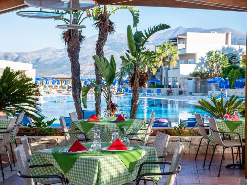 Vista de una piscina con mesas bajo un área cubierta, decorada con palmeras verdes y montañas al fondo.