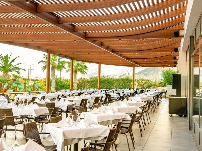 Restaurante al aire libre con filas de mesas y sillas bajo un toldo de madera y palmeras al fondo.