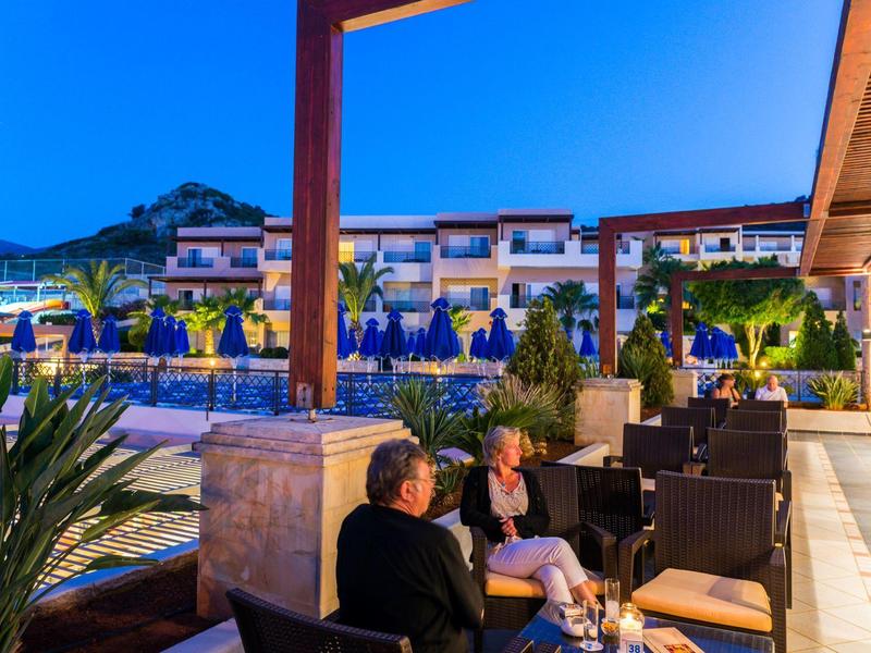 Terraza de hotel con huéspedes al atardecer, zona de asientos con plantas y edificios iluminados de fondo.