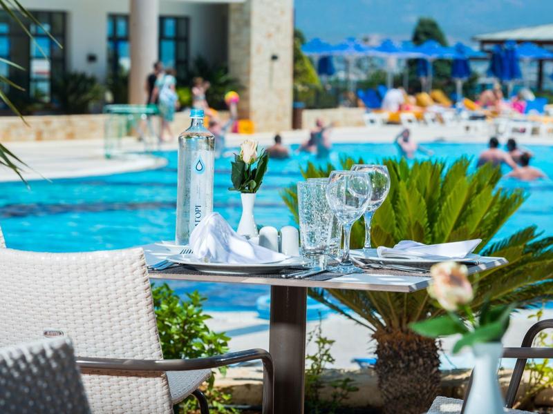 Una mesa puesta con vasos y cubiertos junto a una piscina del hotel con sombrillas y personas.
