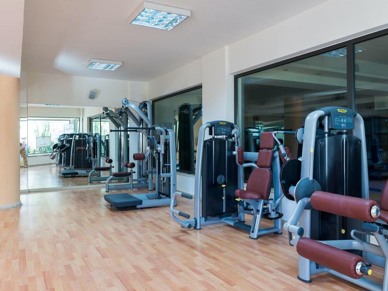 Equipos modernos de gimnasio en una sala amplia y luminosa con suelo de madera y grandes ventanas.