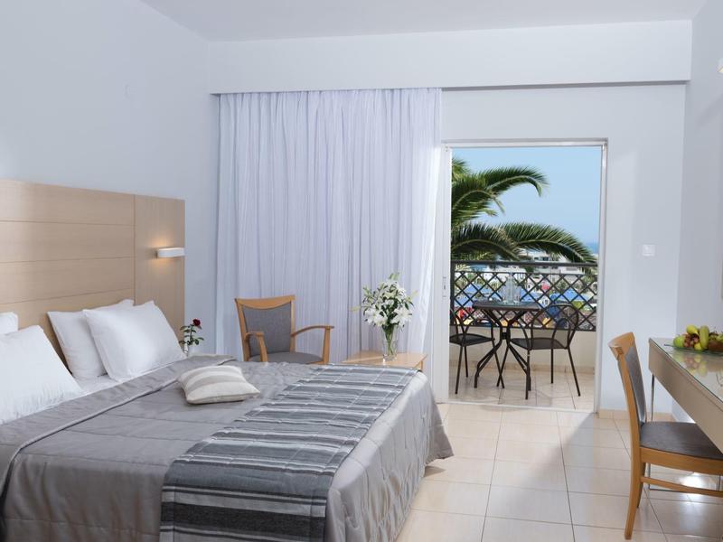 Habitación de hotel moderna con cama doble, escritorio y balcón con vista al mar y palmera tropical.