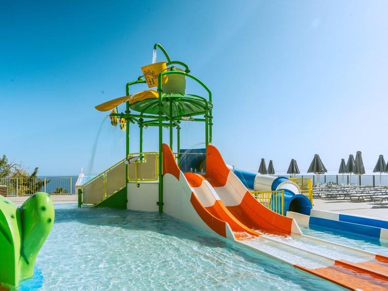 Área de juegos acuáticos para niños con toboganes y agua poco profunda bajo cielo despejado en la piscina del hotel.