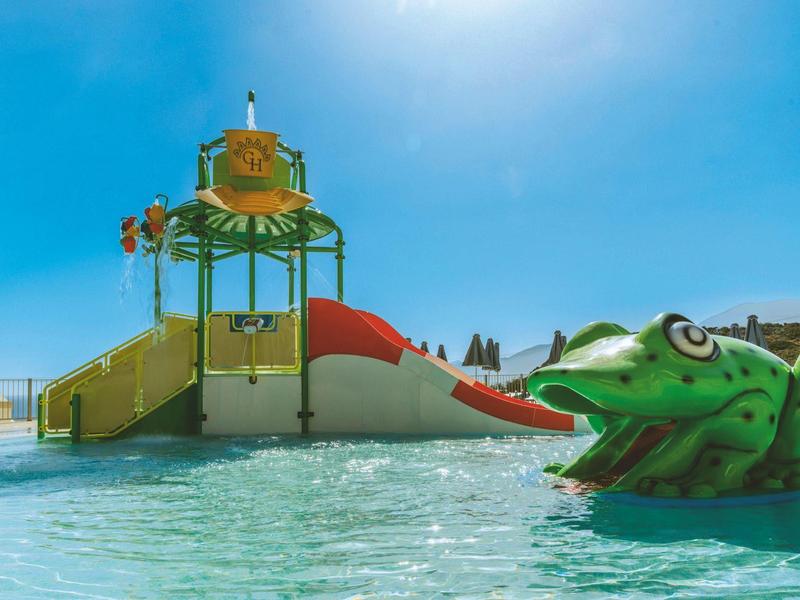 Parque infantil con tobogán acuático y figura de rana en una piscina azul bajo cielo despejado.