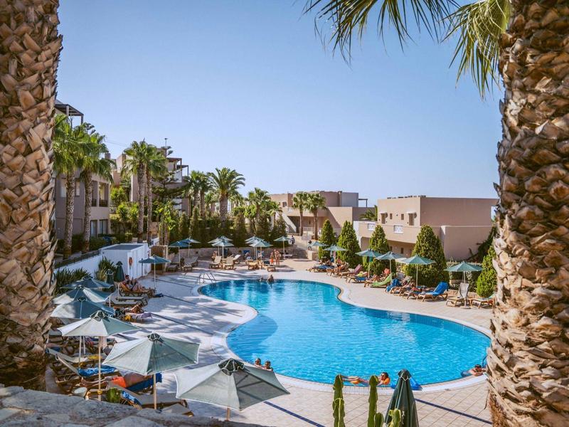 Gran piscina azul en un complejo hotelero con palmeras y sombrillas bajo un cielo despejado.