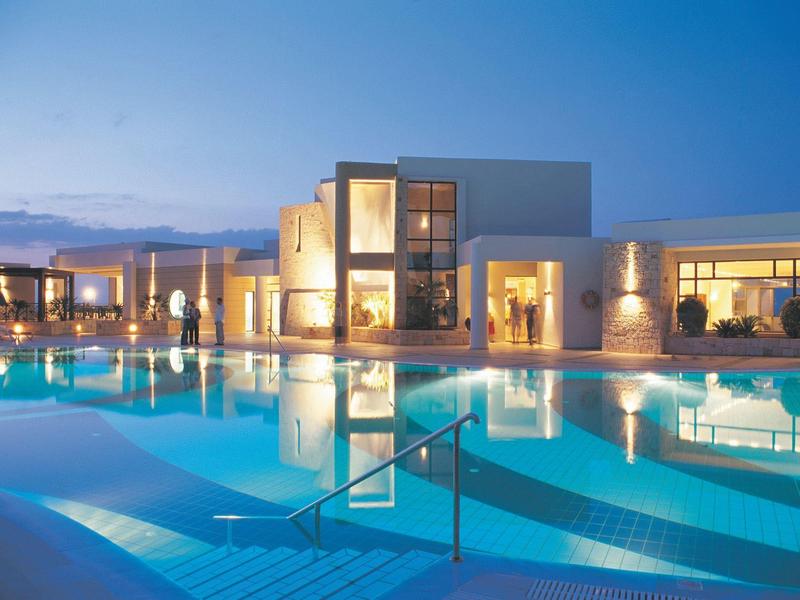 Piscina moderna de hotel con iluminación y edificios al fondo al atardecer.