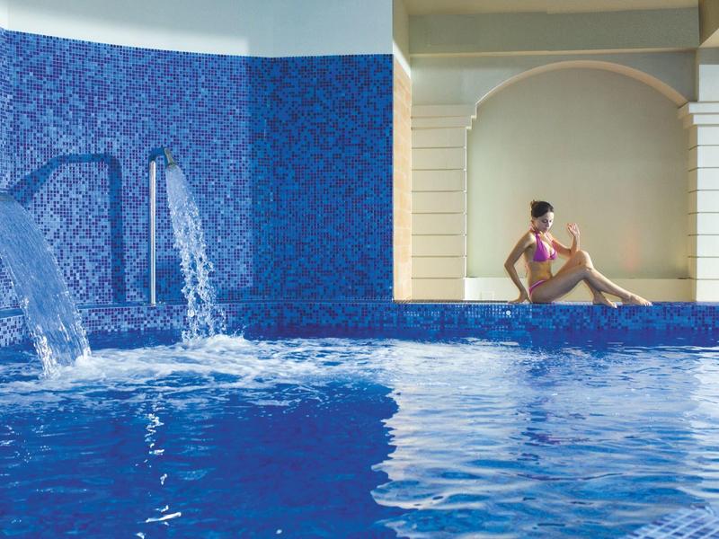 Mujer al borde de una piscina interior azul con cascadas y paredes blancas al fondo.