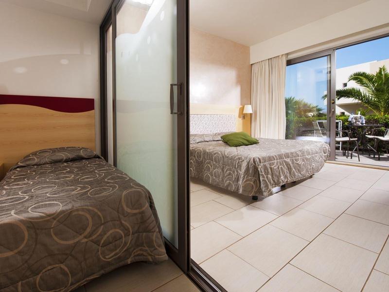 Habitación de hotel con cama individual y doble, puerta de terraza y vista al balcón con plantas.