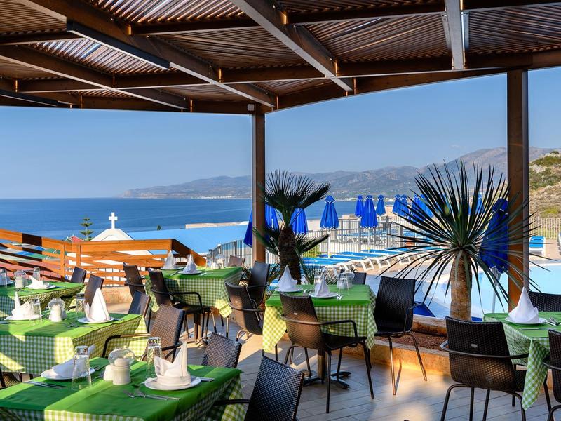 Terraza de restaurante con mesas verdes y vista al mar y tumbonas.