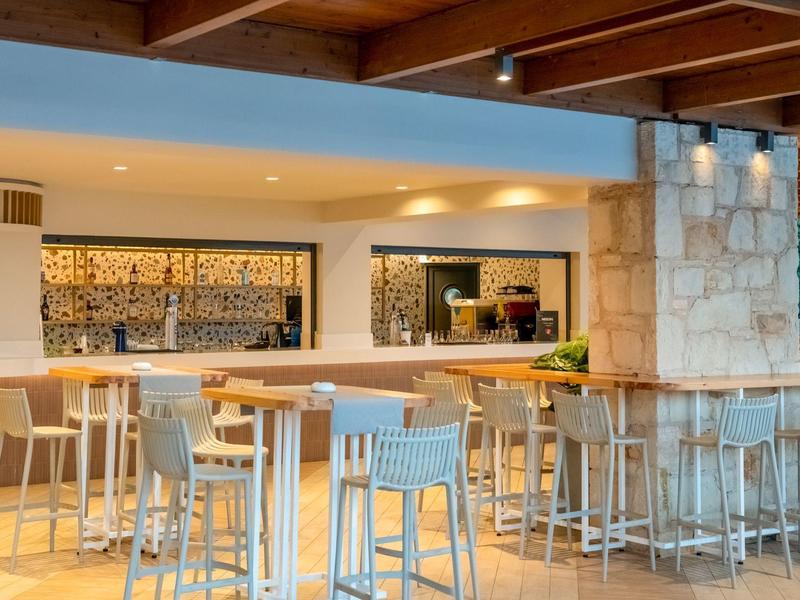 Área de bar moderna y luminosa con vigas de madera, mesas altas y sillas claras.