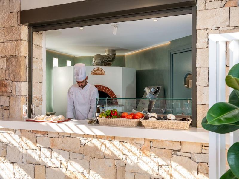 Un chef trabaja detrás de una barra de piedra en una cocina con verduras frescas y horno de pizza.