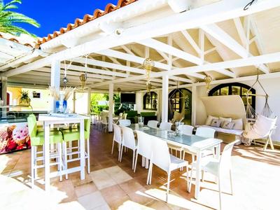 Světlá terasa s jídelním koutem, bílým nábytkem a závěsným křeslem pod střechou s viditelnými trámy.