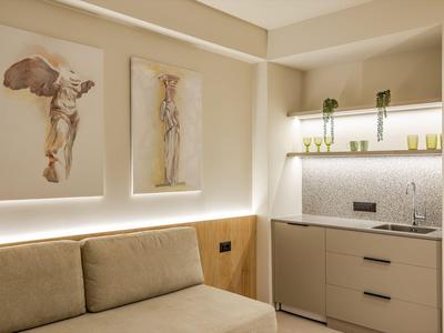 Salón moderno de hotel con sofá beige, dos obras de arte en la pared y pequeña cocina.