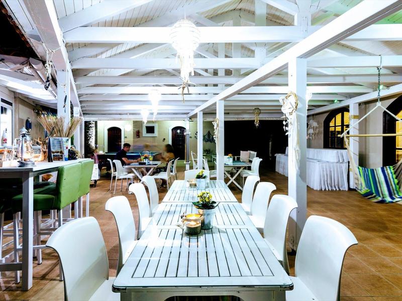 Světlá restaurace s dlouhým dřevěným stolem, bílými židlemi a moderním dekorem