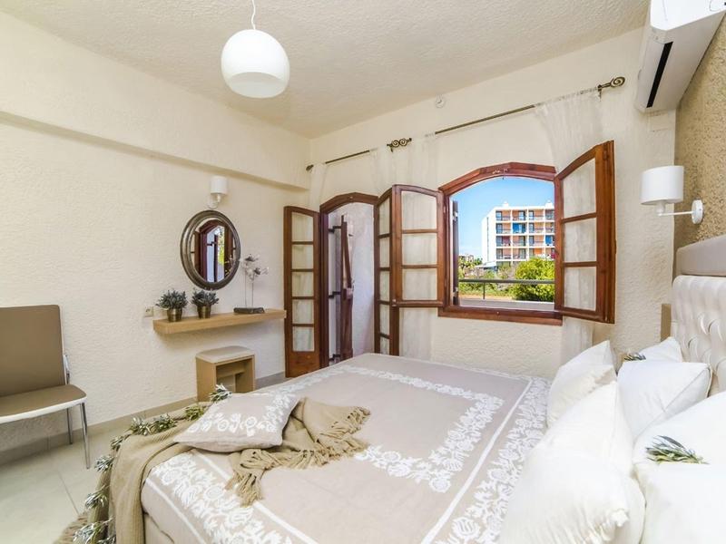 Světý hotelový pokoj s manželskou postelí, stolem, židlí a otevřeným oknem s výhledem na budovy.