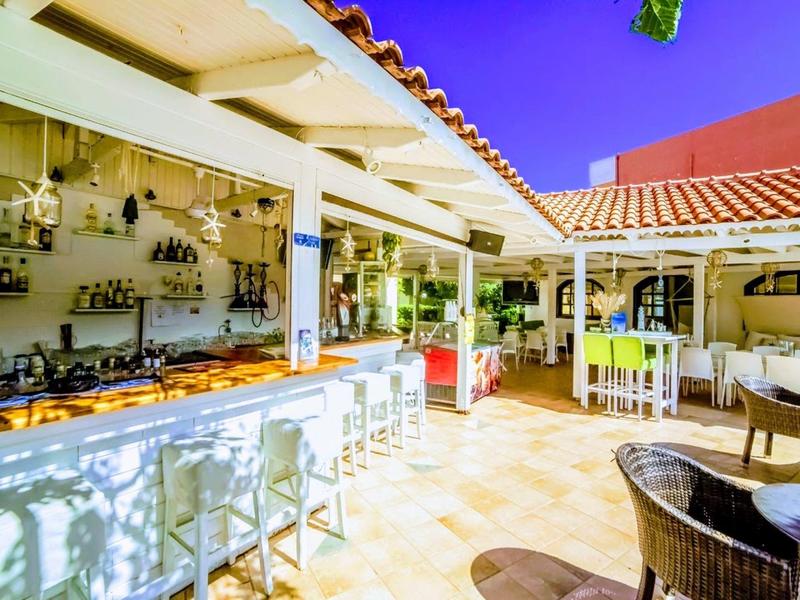 Světlý venkovní bar s vysokým pultem, stoličkami a posezením pod zastřešenou střechou.