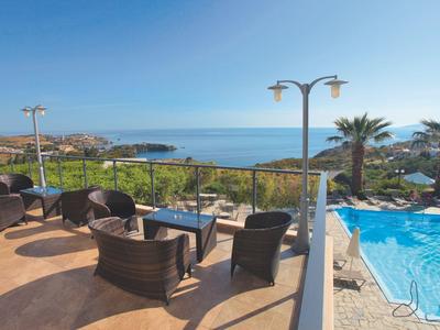Terrasse mit schwarzen Sitzmöbeln, Blick auf Meer, Pool und Palmen unter klarem blauem Himmel.