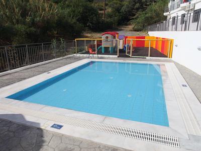 Rechteckiger blauer Pool mit weißem Rand, umgeben von einer grauen Steinfläche und buntem Zaun.