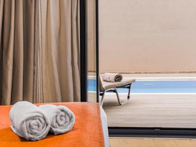 Zwei weiße Handtücher auf orangefarbenem Bett vor Glasfenster mit Blick auf Pool und Liegestuhl.