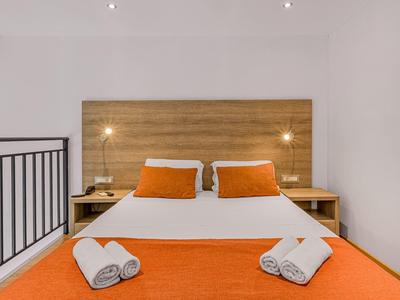 Modernes Schlafzimmer mit Doppelbett, orange Decke, zwei Kissen und je zwei gerollten Handtüchern.