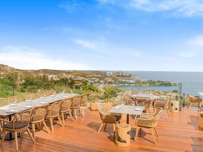 Střešní terasa s dřevěnou podlahou, židlemi a stoly s výhledem na moře a pobřeží.