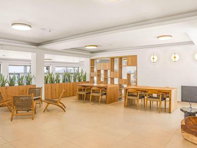 Světlá společná místnost s dřevěnými židlemi, stoly a velkými okny s rostlinami.