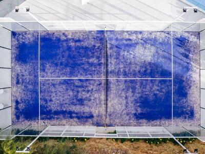 Letecký pohled na obdélníkové sportovní hřiště s modrým kobercem, obklopené plotem.