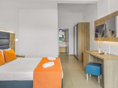 Moderní hotelový pokoj s dvojlůžkem, oranžovými akcenty a zrcadlem nad psacím stolem.