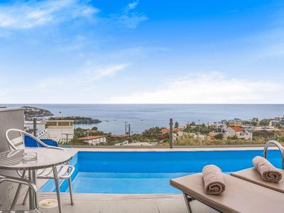 Bazén s lehátky a židlemi s výhledem na moře a město pod modrou oblohou.