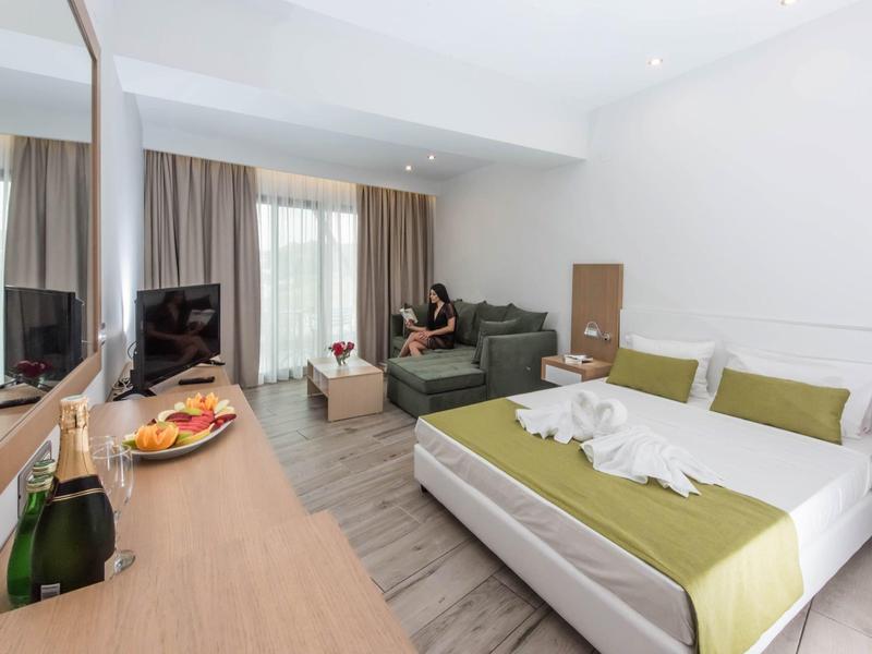 Moderně zařízený hotelový pokoj s manželskou postelí, pohovkou, televizí a talířem ovoce na stole.