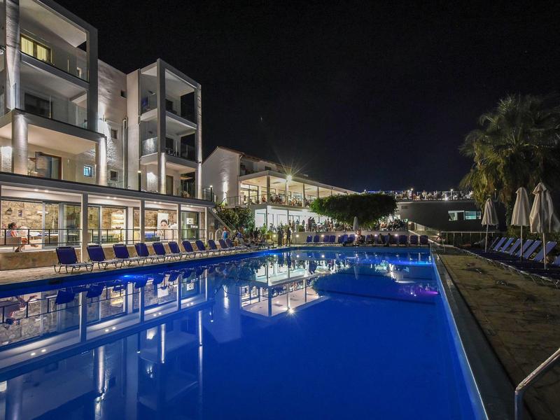 Noční pohled na hotel s osvětleným bazénem a lehátky po stranách.