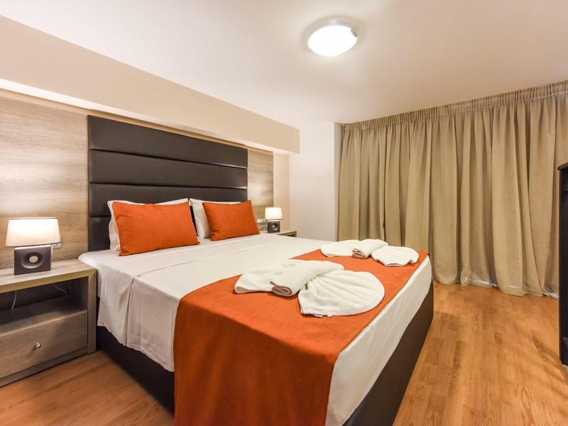 Moderně zařízený hotelový pokoj s manželskou postelí, oranžovými detaily a dřevěnou podlahou.