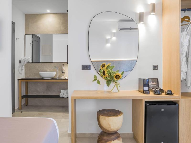 Světlý hotelový pokoj s umyvadlem, zrcadlem, stolem se slunečnicemi a mini ledničkou.