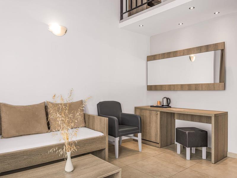 Moderně zařízený hotelový pokoj s pohovkou, křeslem, psacím stolem a velkým zrcadlem.