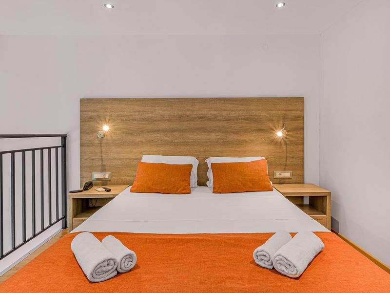 Moderně zařízený hotelový pokoj s manželskou postelí, oranžovým přehozem a bílými ručníky.