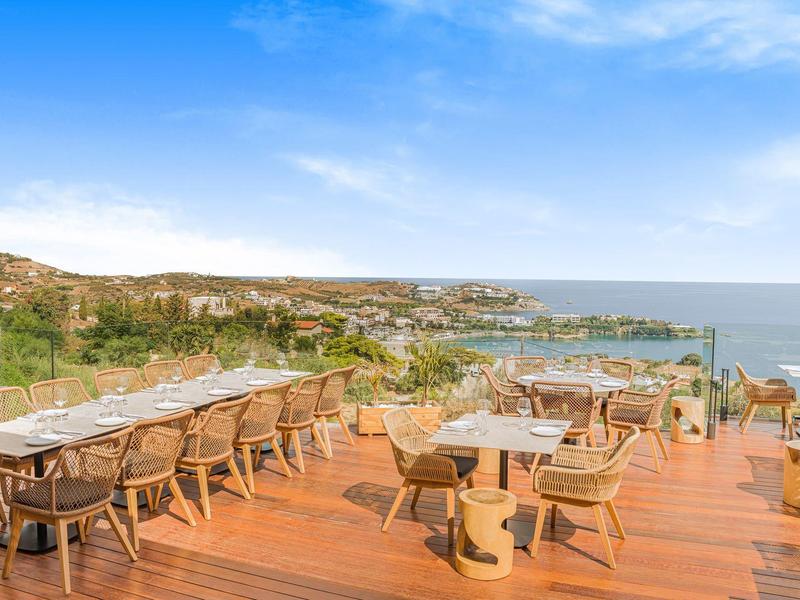 Střešní terasa s dřevěnou podlahou, židlemi a stoly s výhledem na moře a pobřeží.