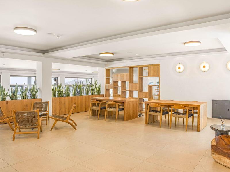 Světlá společná místnost s dřevěnými židlemi, stoly a velkými okny s rostlinami.