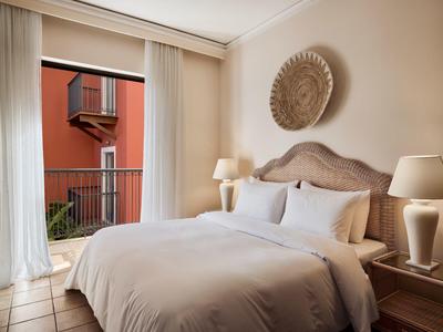 Helles Schlafzimmer mit großem Bett, weißen Vorhängen, zwei Nachttischlampen und Tür zum Balkon.