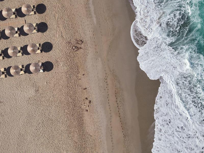 Luftaufnahme eines Strandes mit Liegestühlen und Sonnenschirmen neben brechenden Wellen