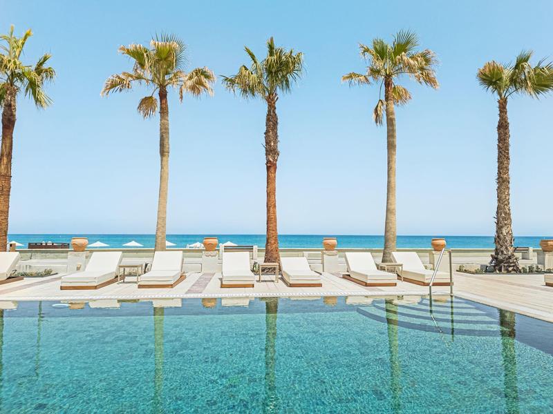 Schwimmbad mit Liegen und Palmen neben dem Meer bei klarem Himmel.