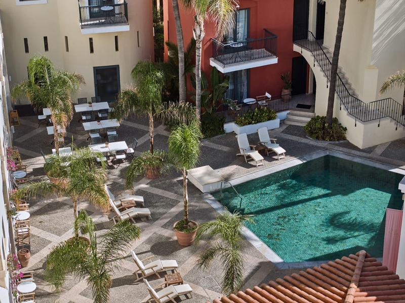 Hotelpool mit Liegestühlen und Palmen in einem Innenhof mit Terrassen und roten Wänden.