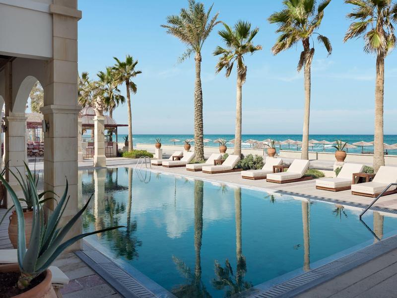 Moderner Pool mit Palmen und Liegen am Meer unter klarem blauem Himmel.