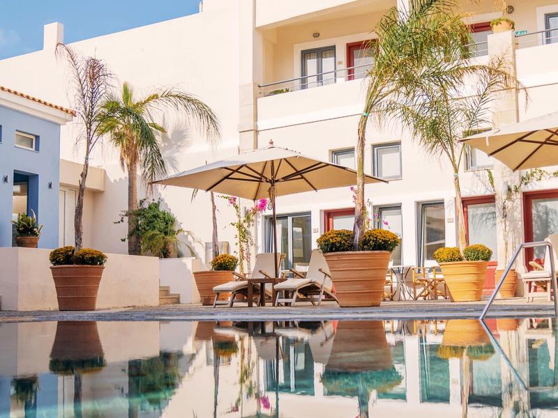 Moderner Hotelpoolbereich mit Sonnenschirmen, Palmen und weißen Gebäuden im Hintergrund.