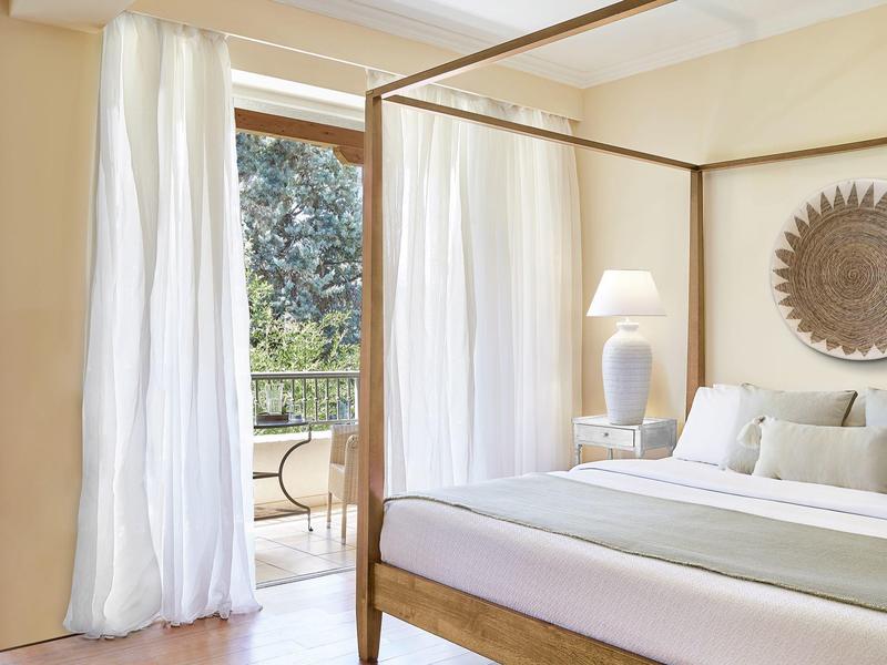 Helles Hotelzimmer mit Himmelbett, weißer Bettwäsche, Lampe und Zugang zu Balkon mit Bäumen.